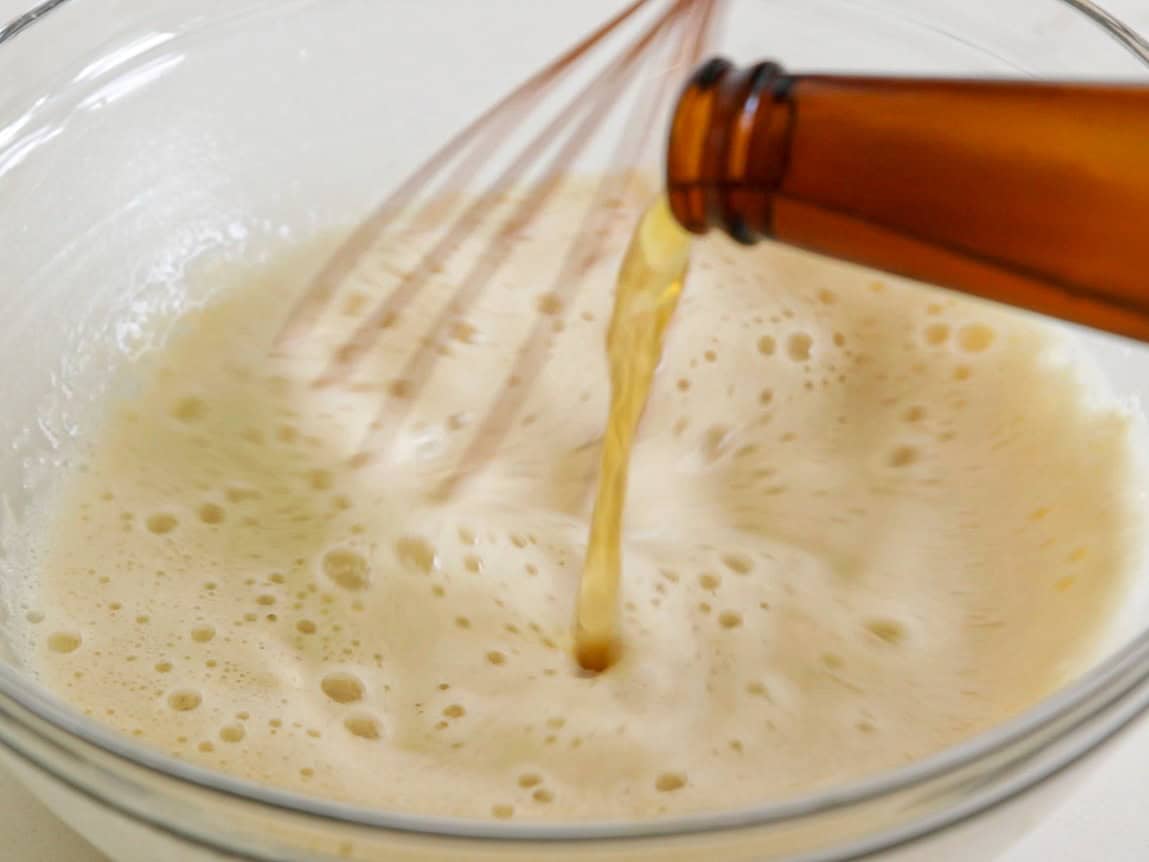 A close-up of a brown bottle pouring beer into a glass bowl with a foamy mixture. A whisk is partially visible, suggesting an ingredient is being mixed in preparation for a recipe.
