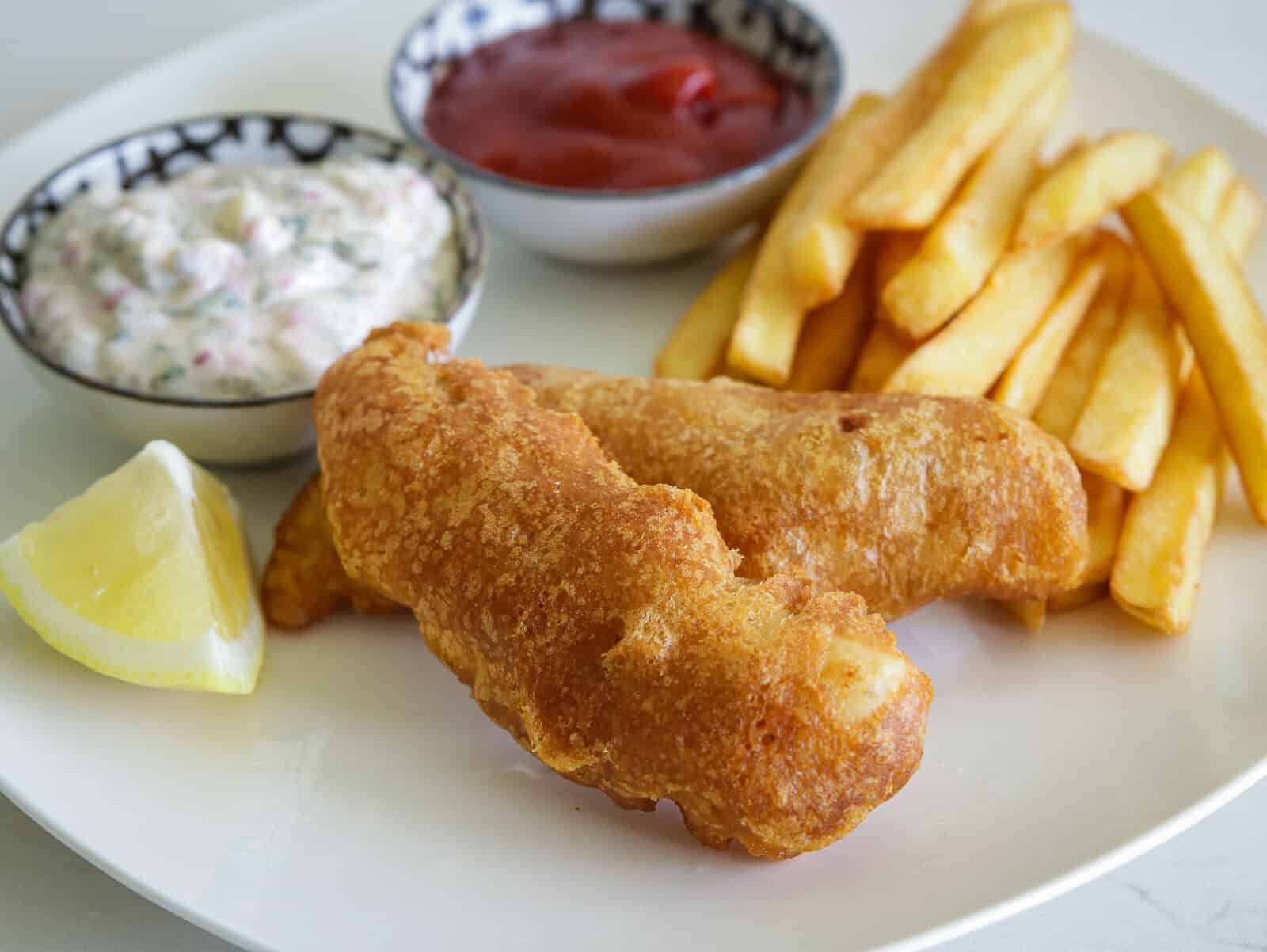 A plate with two pieces of golden fried beer battered fish and chips, thick-cut fries, a lemon wedge, and small bowls of tartar sauce and ketchup.