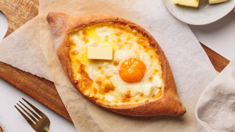 A baked khachapuri bread filled with melted cheese, topped with a sunny-side-up egg yolk and a pat of butter, sits on parchment paper beside a fork and a plate with more butter.