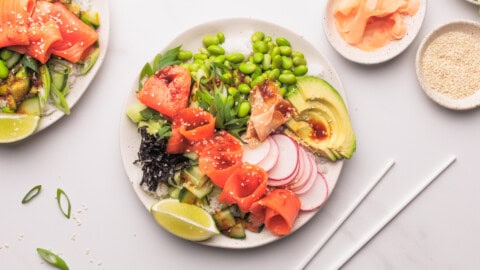 A colorful poke bowl with smoked salmon, avocado, edamame, radish slices, cucumber, seaweed, lime wedges, sesame seeds, and green onions, served with chopsticks, pickled ginger, and a small dish of sesame seeds on the side.