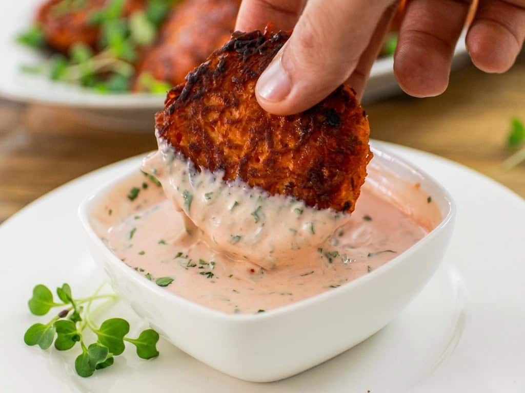 A hand dipping a crispy, golden-brown sweet potato fritter into a creamy, herb-speckled dipping sauce in a white bowl, with microgreens on the plate nearby.