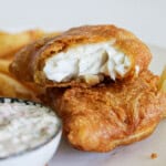 Two pieces of golden-brown battered fish and chips with flaky white interior, stacked on a plate next to thick-cut fries and a small bowl of creamy tartar sauce.