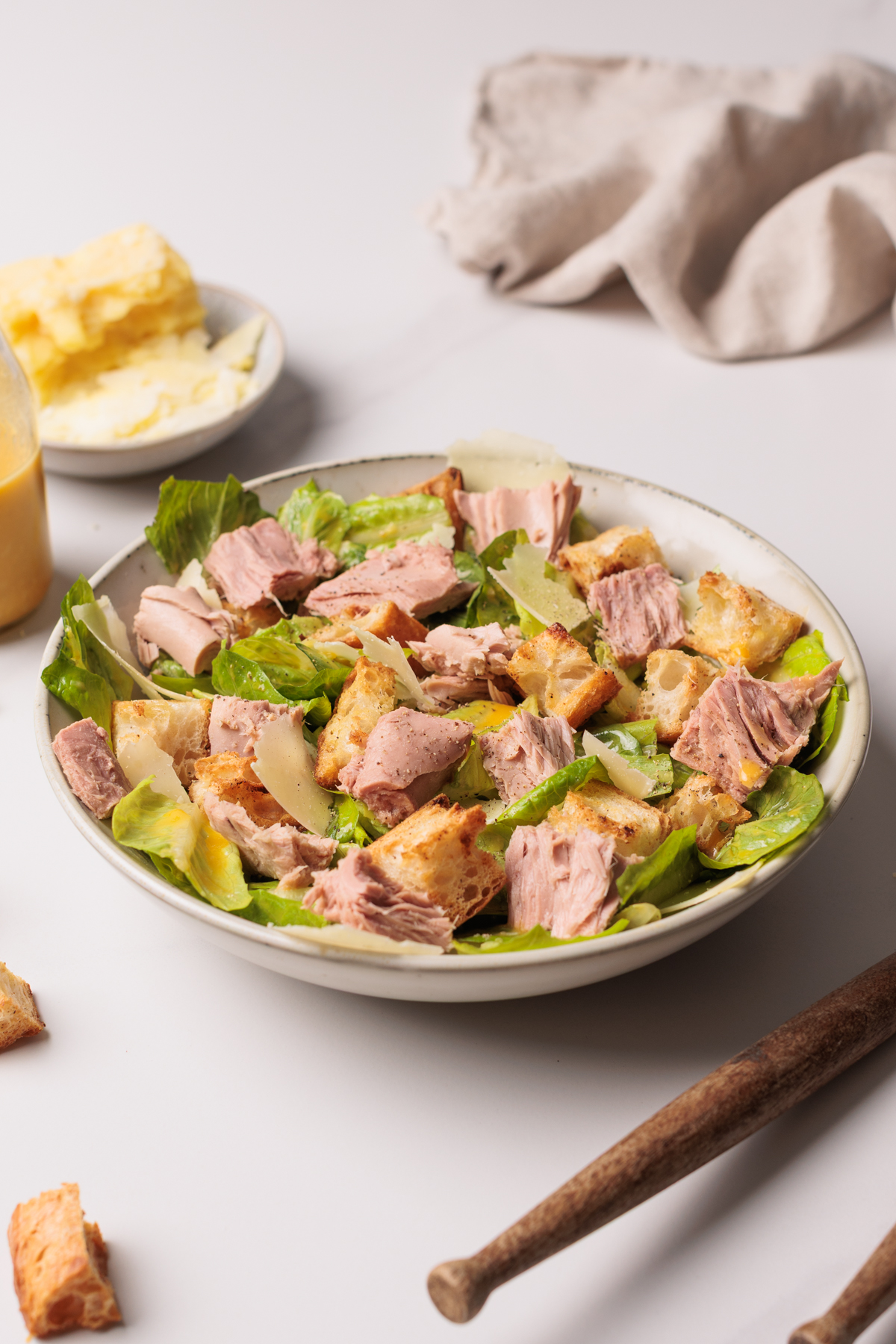 A bowl of salad with chunks of tuna, lettuce, croutons, and cheese on a white surface. A bottle of dressing, a plate with butter, and a beige cloth are in the background.