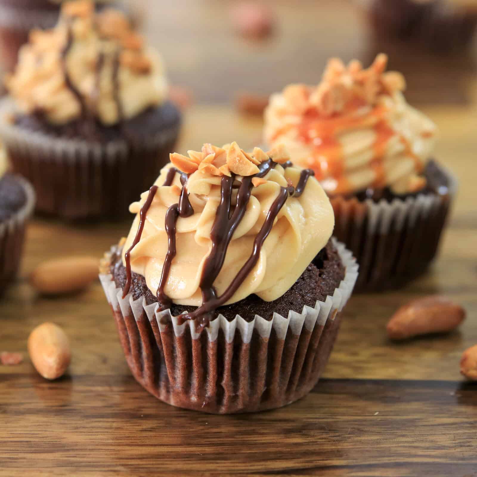 Chocolate cupcakes topped with creamy peanut butter frosting, drizzled with chocolate and caramel sauces, and sprinkled with chopped peanuts, displayed on a wooden surface.