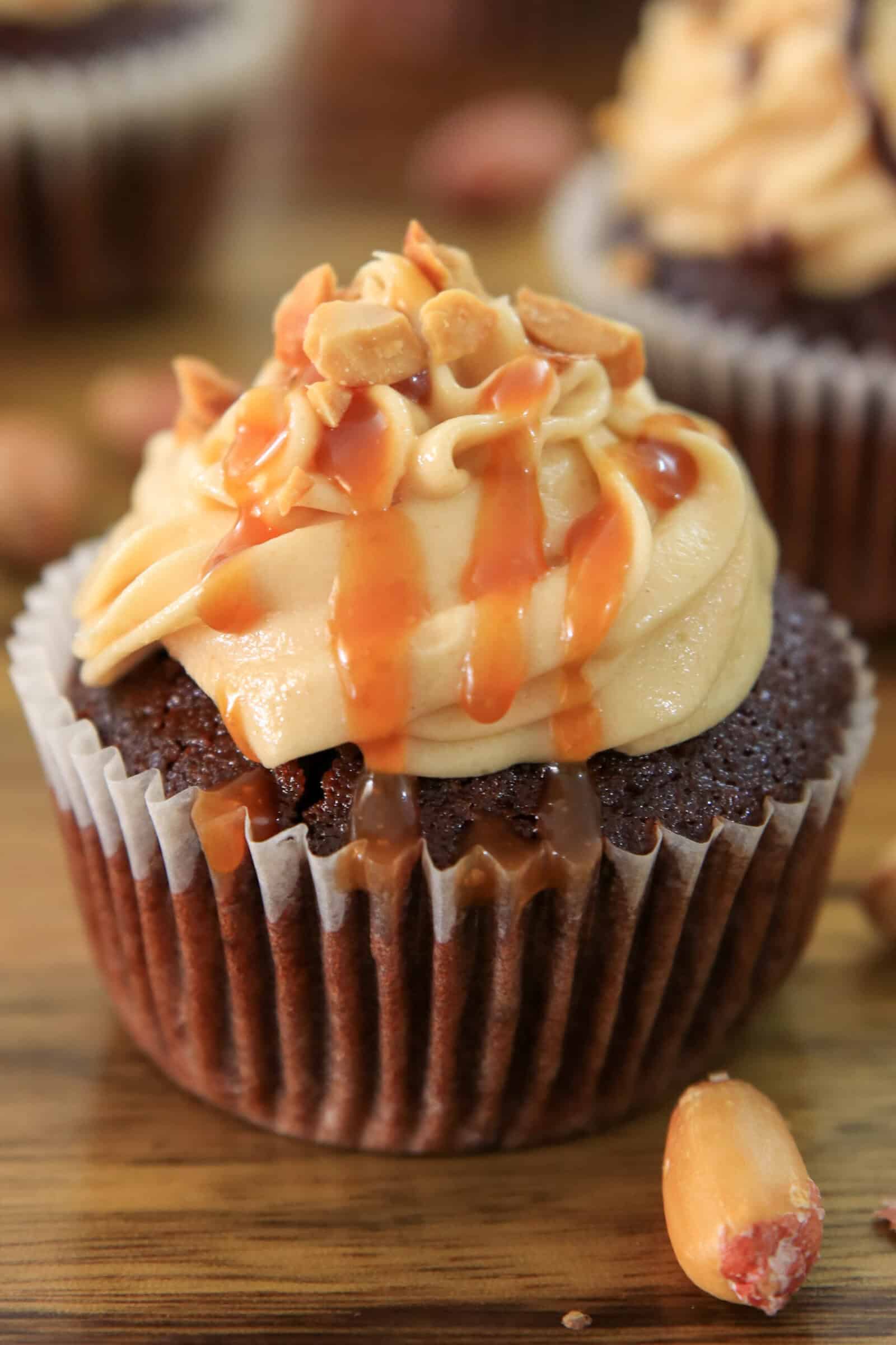 A chocolate peanut butter cupcake with creamy peanut butter frosting, topped with chopped peanuts and drizzled with caramel sauce, sits on a wooden surface with a few peanuts scattered nearby.