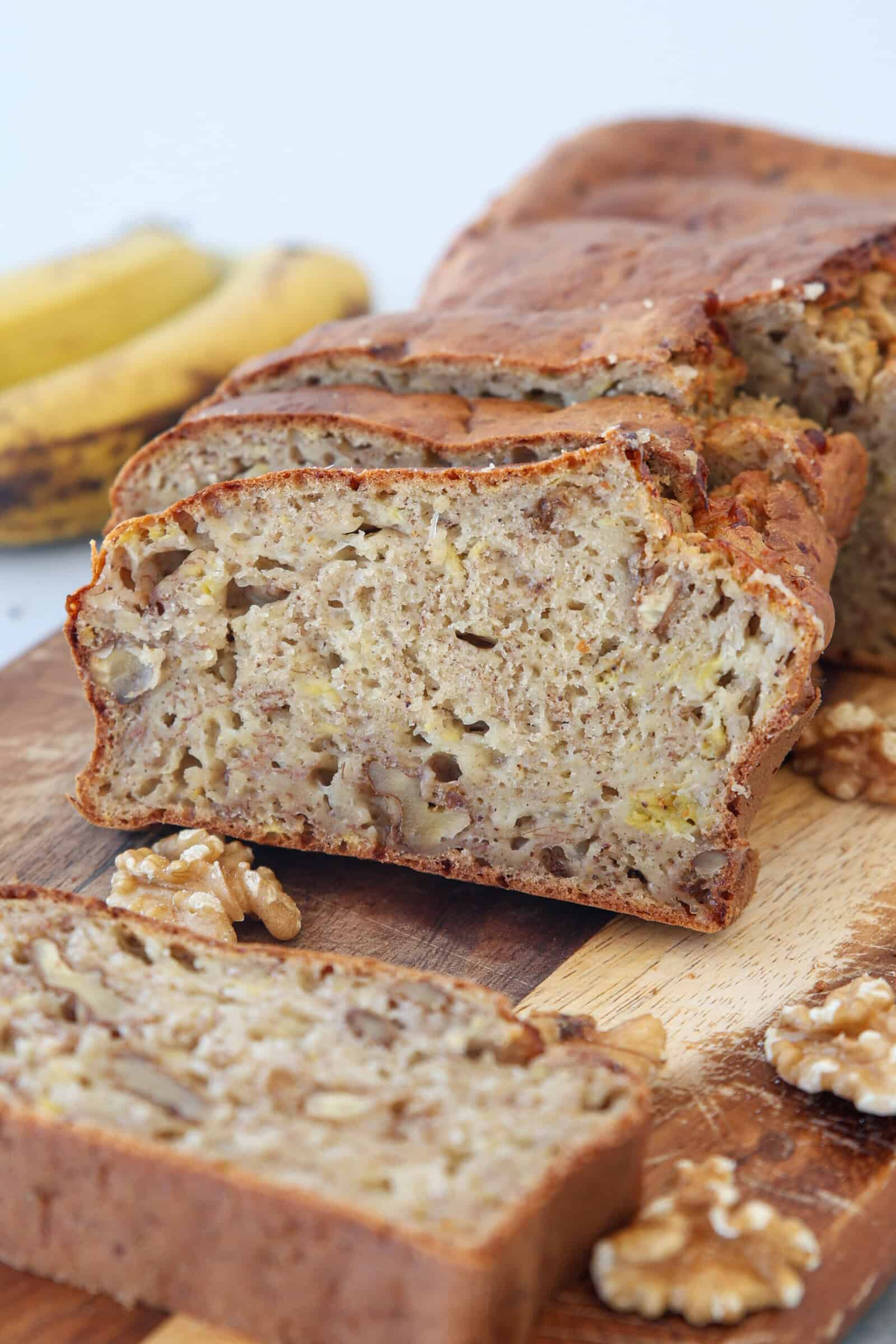 A sliced loaf of cottage cheese banana bread with visible walnuts, placed on a wooden cutting board. Whole walnuts and ripe bananas are also visible in the background.