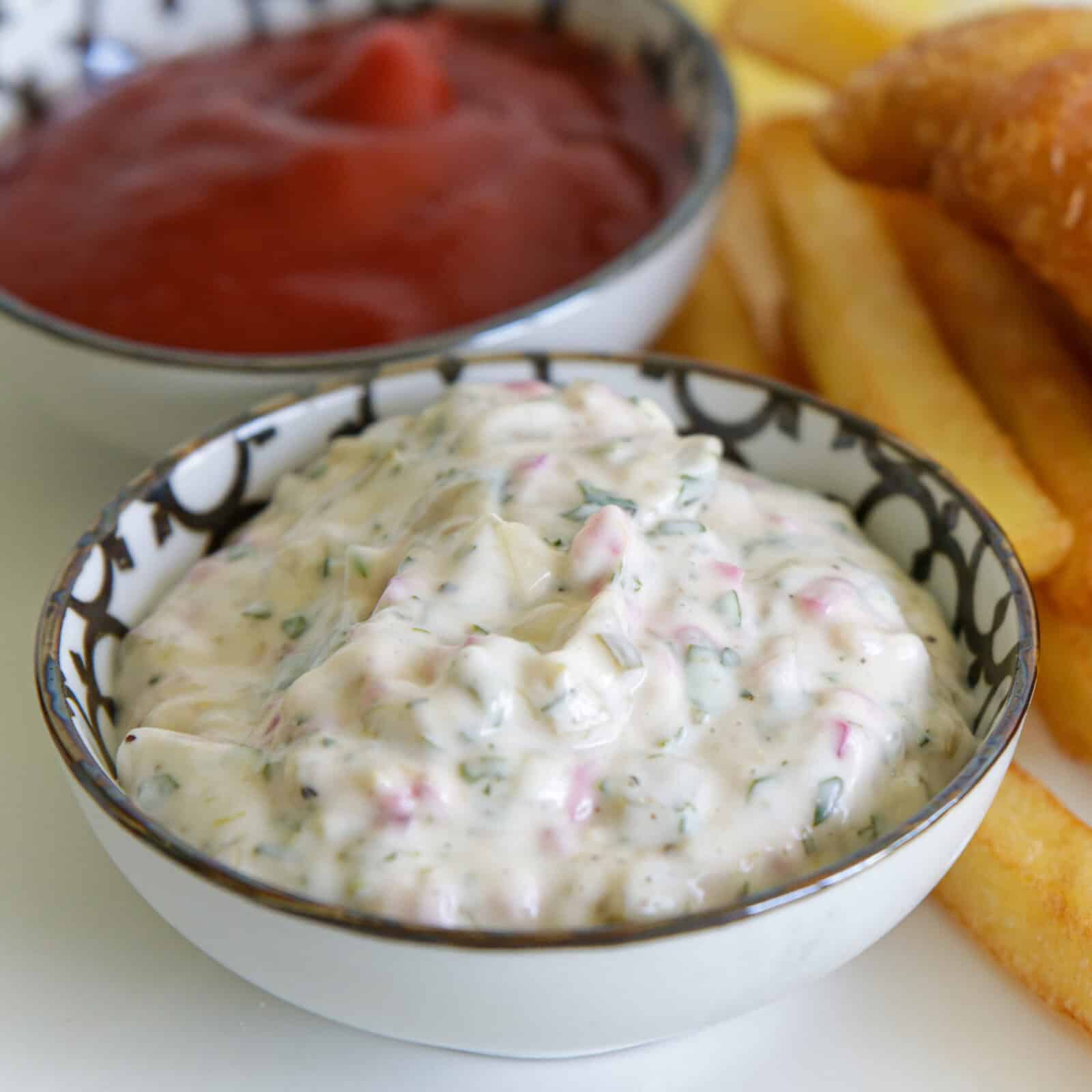 A bowl of creamy tartar sauce with herbs and chopped pickles, next to a bowl of ketchup and a serving of golden French fries.