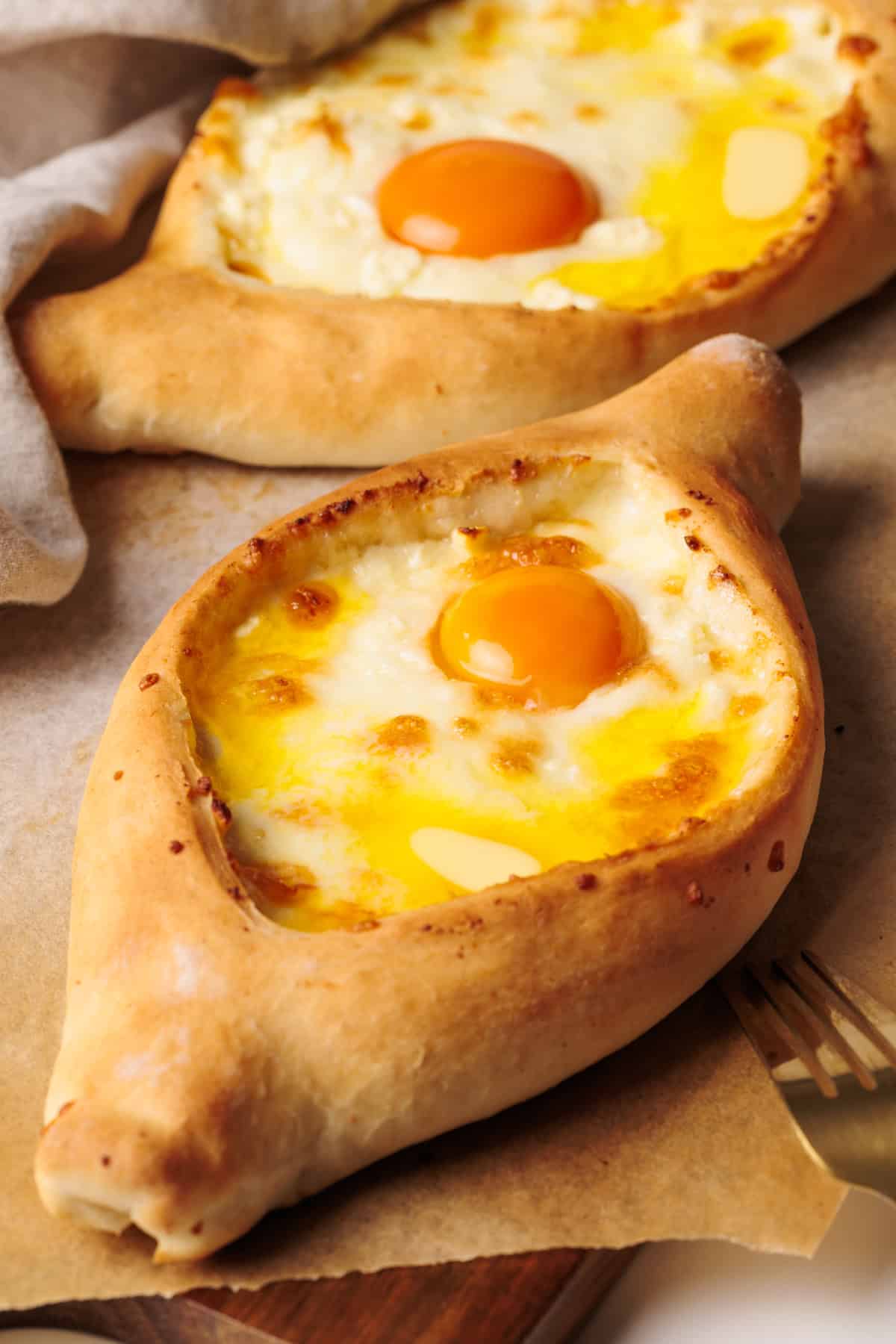 Two khachapuri breads filled with melted cheese and topped with a sunny-side-up egg and small pieces of butter, served on parchment paper with a fork partially visible beside them.