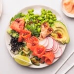A bowl filled with white rice, sliced smoked salmon, avocado, edamame, cucumber, radish, seaweed, green onions, lime wedge, and a drizzle of sauce, garnished with sesame seeds.