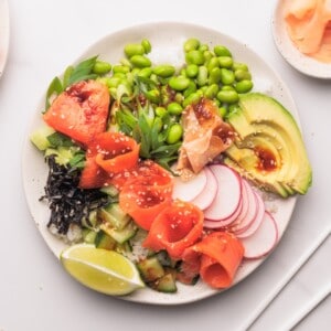 A bowl filled with white rice, sliced smoked salmon, avocado, edamame, cucumber, radish, seaweed, green onions, lime wedge, and a drizzle of sauce, garnished with sesame seeds.