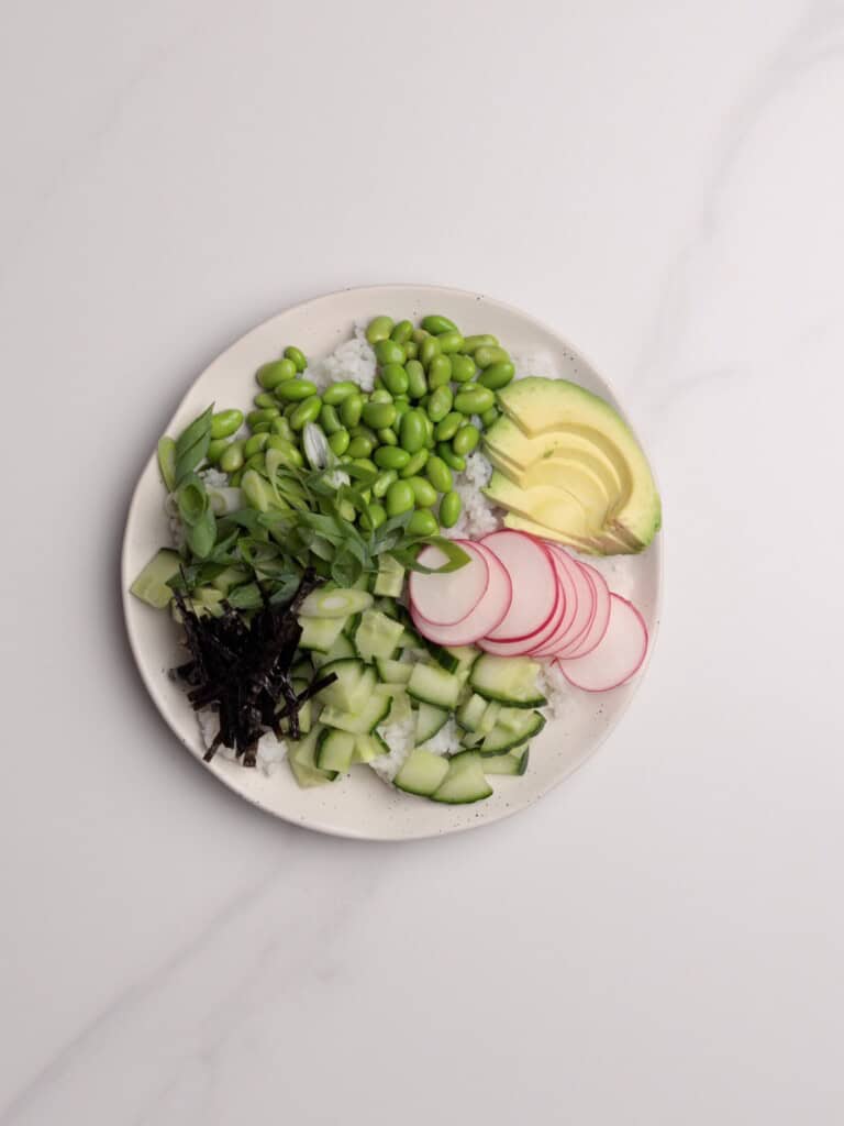 A plate with sliced avocado, radishes, cucumber, green onions, edamame, nori strips, and rice arranged in separate sections on a light surface.