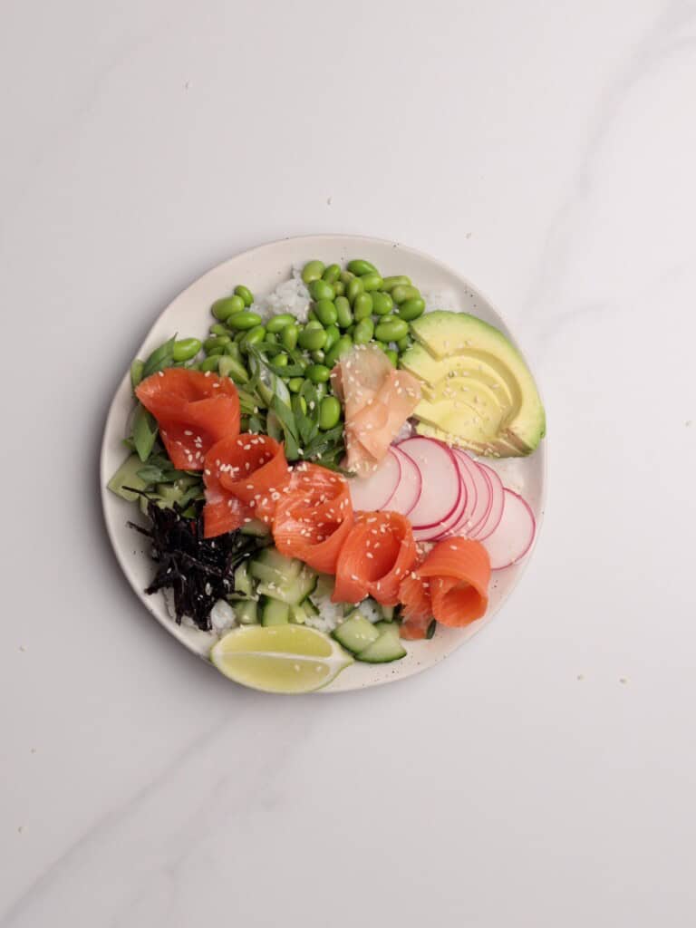 A bowl with sliced avocado, radish, cucumber, seaweed, edamame, pickled ginger, lemon wedge, and salmon roses, garnished with sesame seeds, on a white marble surface.