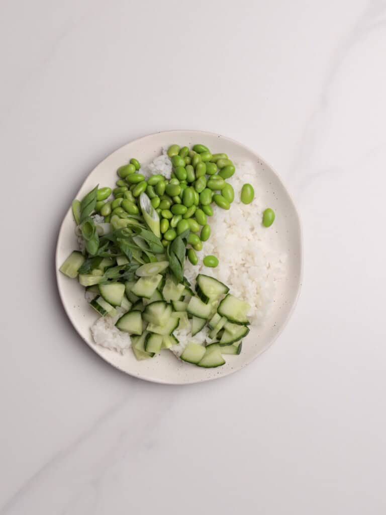 A plate with sections of white rice, chopped cucumber, sliced green onions, and green edamame beans, arranged separately on a white marble surface.