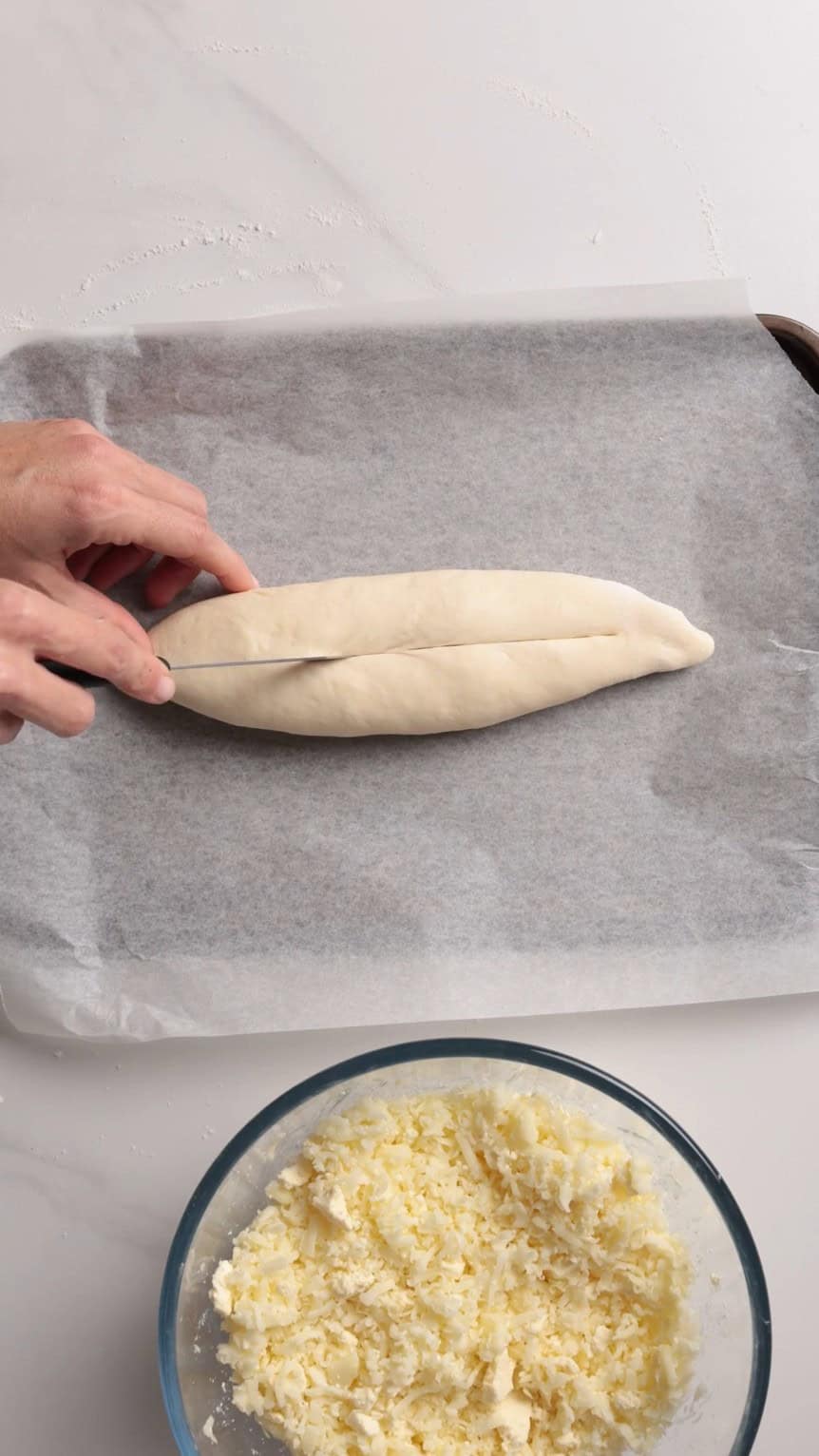 A hand slices a loaf-shaped dough on parchment paper, preparing it for baking. A glass bowl filled with grated cheese sits nearby on a white countertop.