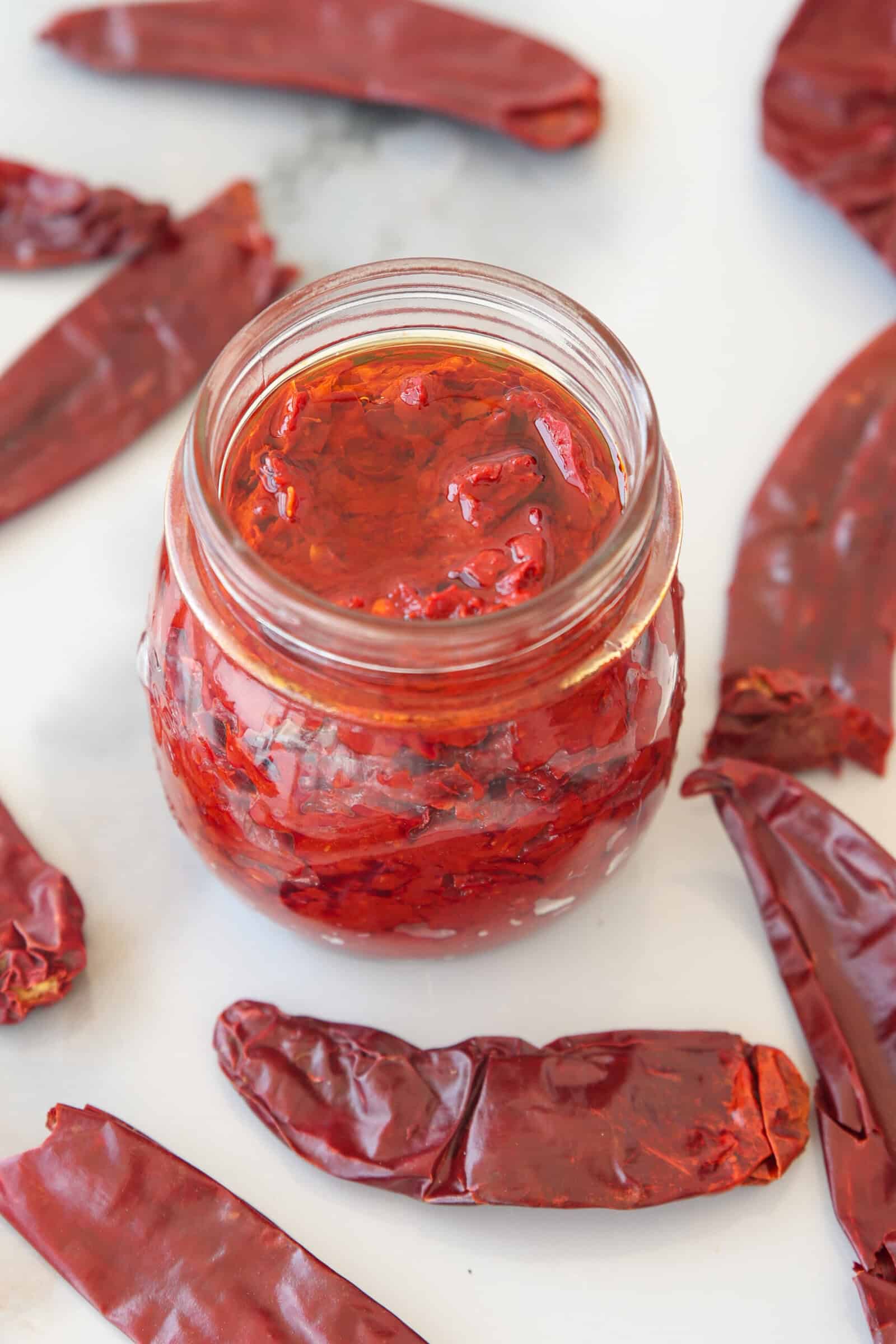 A glass jar filled with harissa and pieces of chili sits on a white surface, surrounded by several whole dried red chilies.