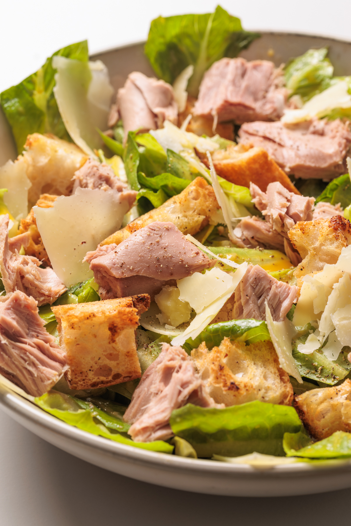A close-up of a Tuna Caesar Salad featuring chunks of tuna, romaine lettuce, shaved Parmesan cheese, and crispy croutons in a white bowl.