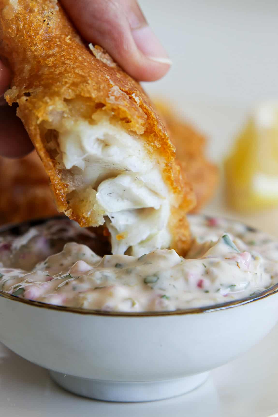 A hand dips a piece of crispy fried fish with flaky white inside into a bowl of creamy tartar sauce with visible herbs and relish. A lemon wedge is blurred in the background.