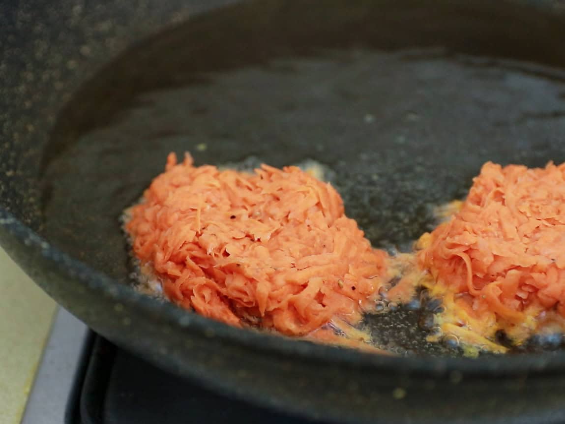 Two raw, shredded sweet potato patties are frying in oil in a black skillet on a stovetop.
