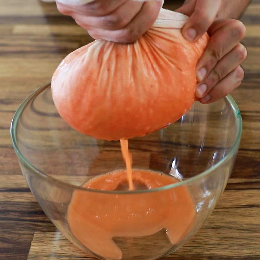 A person squeezes an orange liquid through a cheesecloth or nut milk bag over a clear glass bowl, extracting juice or blended mixture onto a wooden surface.