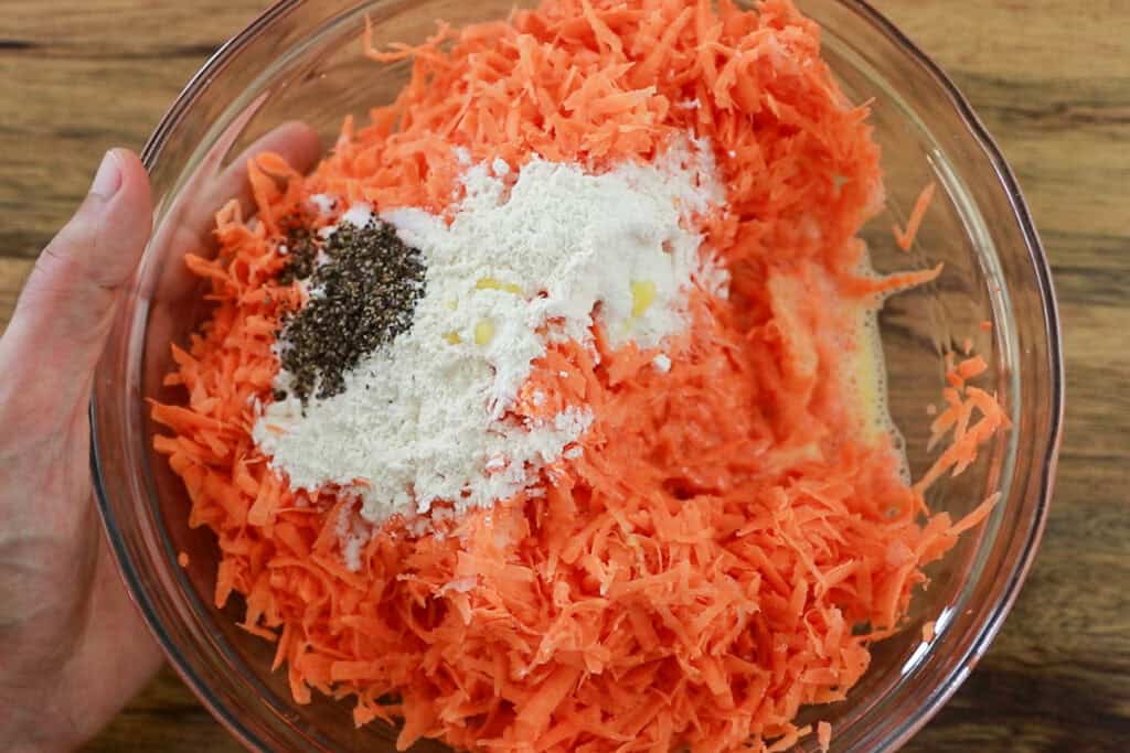 A glass bowl filled with grated sweet potato, flour, black pepper, and other ingredients, held in one hand over a wooden surface, ready to be mixed for cooking or baking.