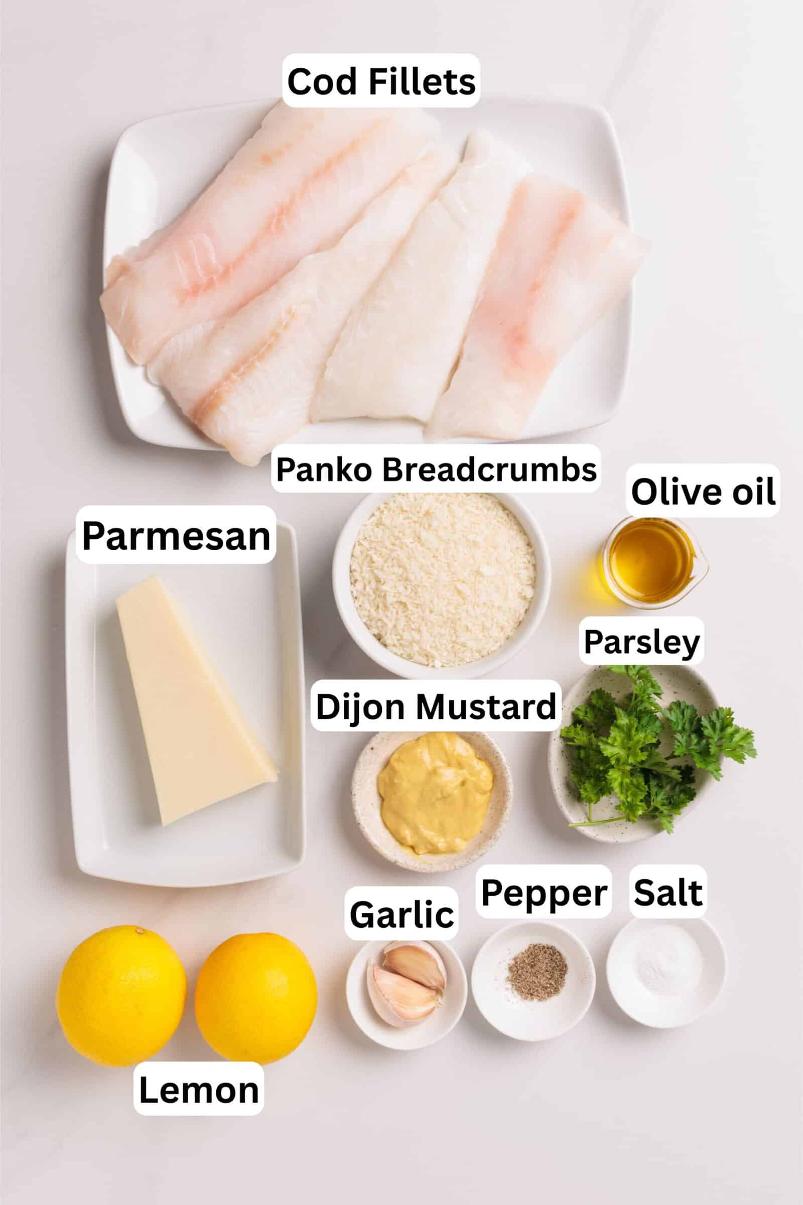 Overhead view of labeled ingredients for a recipe: three cod fillets, panko breadcrumbs, olive oil, parsley, Parmesan cheese, Dijon mustard, garlic cloves, lemon, salt, and pepper.