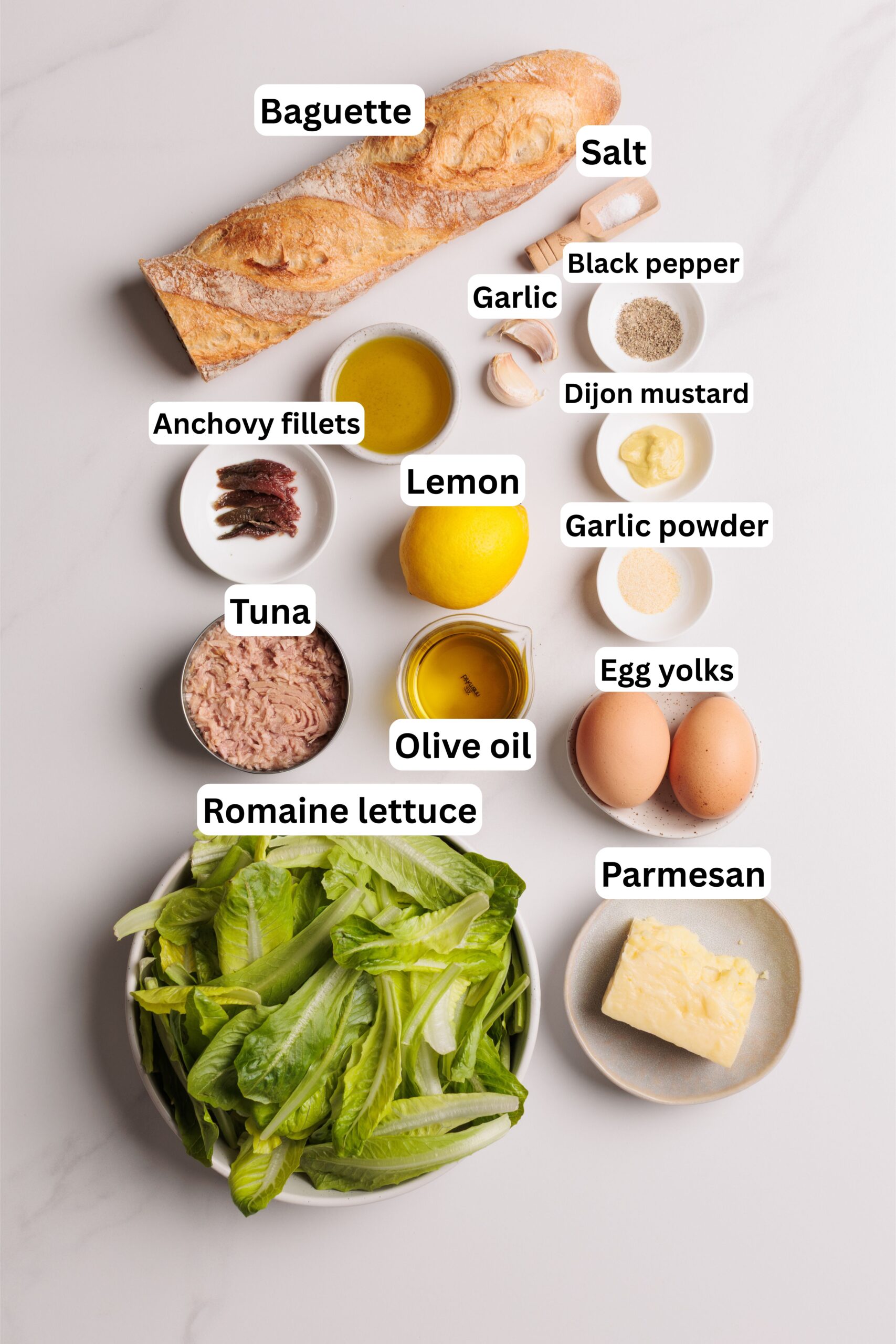 A flat lay of Caesar salad ingredients: baguette, salt, black pepper, garlic, anchovy fillets, tuna, lemon, Dijon mustard, garlic powder, olive oil, egg yolks, romaine lettuce, and Parmesan cheese.