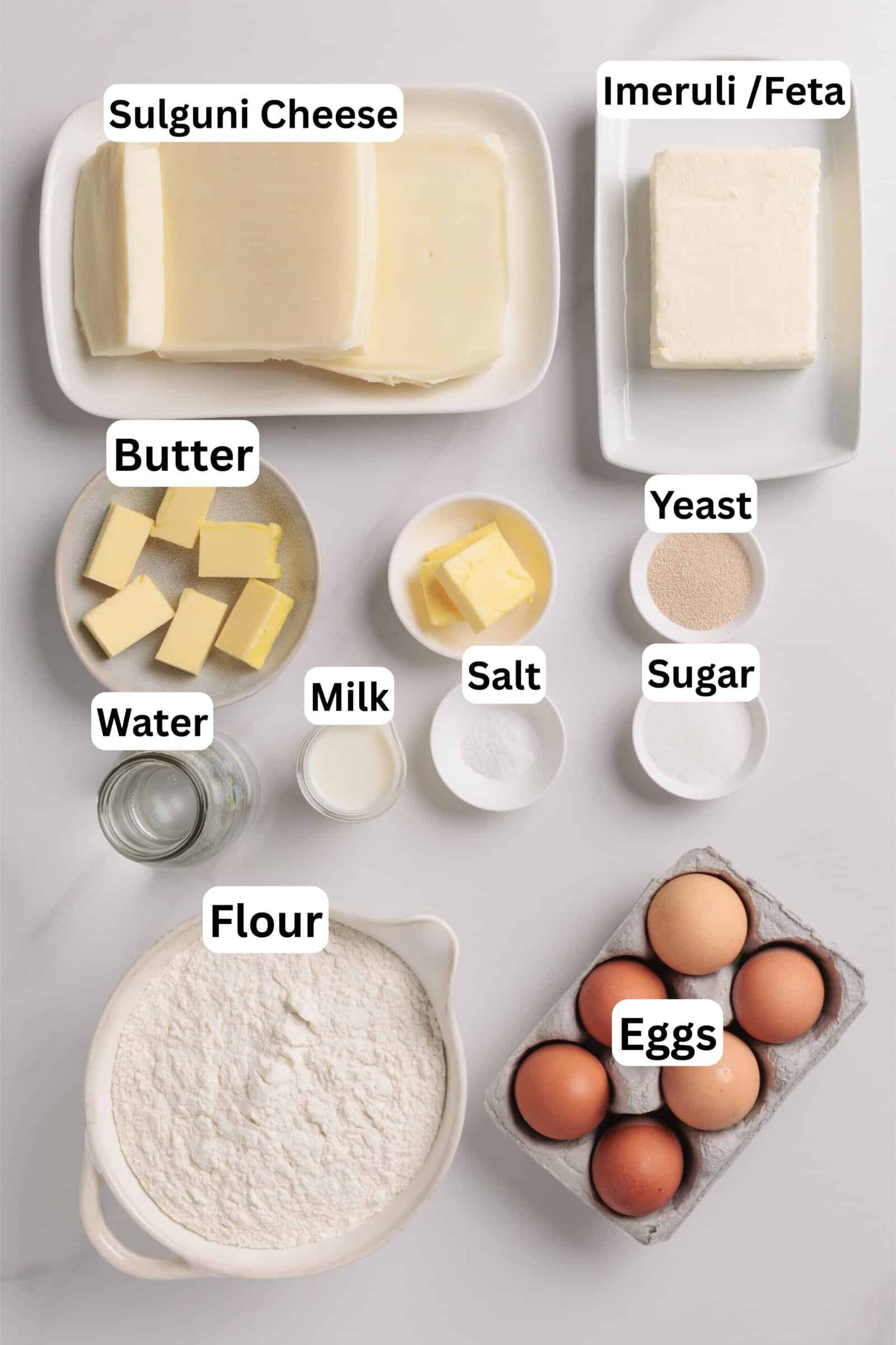 Top-down view of ingredients for bread or pastry: sliced sulguni cheese, block of Imeruli/feta, butter, yeast, sugar, salt, milk, water, flour, and a carton of brown eggs, each labeled.
