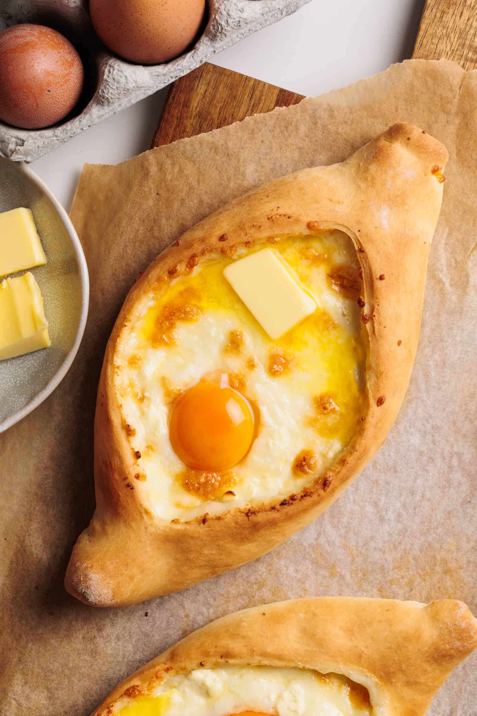 A freshly baked khachapuri bread filled with melted cheese, a sunny-side-up egg, and a pat of butter on top, placed on parchment paper with eggs and butter nearby.