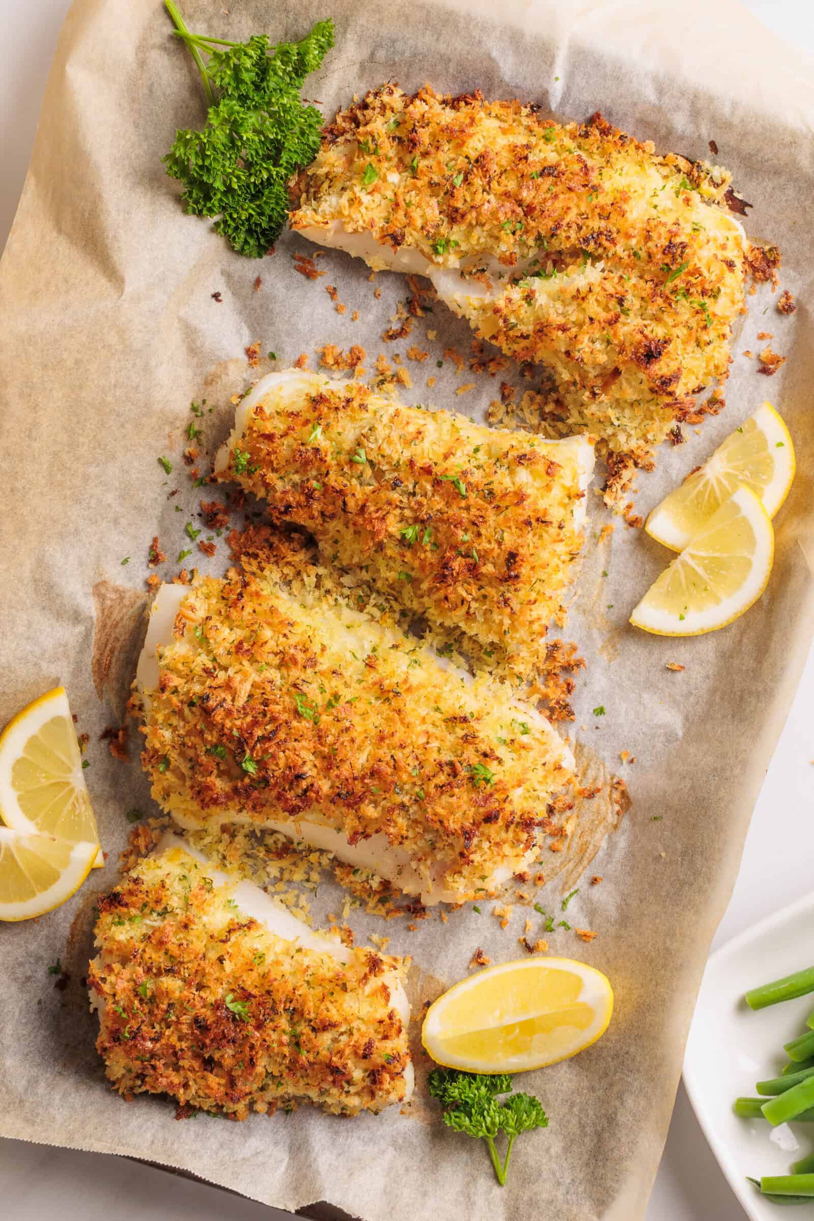 Four pieces of baked cod fish with a golden, crispy panko breadcrumb topping on parchment paper, garnished with parsley and served with lemon wedges.