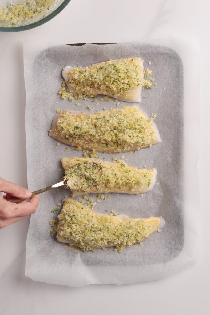 A hand spreads apanko breadcrumb and herb mixture onto raw fish fillets arranged on a parchment-lined baking sheet.