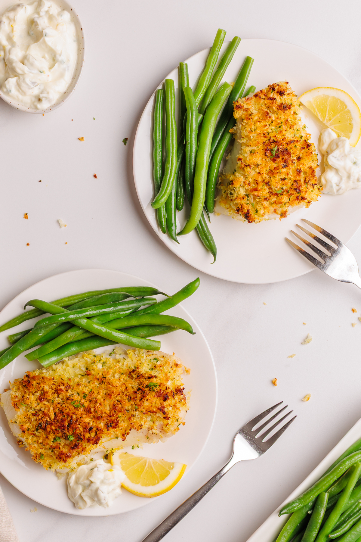 Two plates with crispy baked Parmesan-crusted cod fish, green beans, lemon wedges, and a dollop of tartar sauce, with a fork next to each plate and a small bowl of tartar sauce nearby on a light surface.