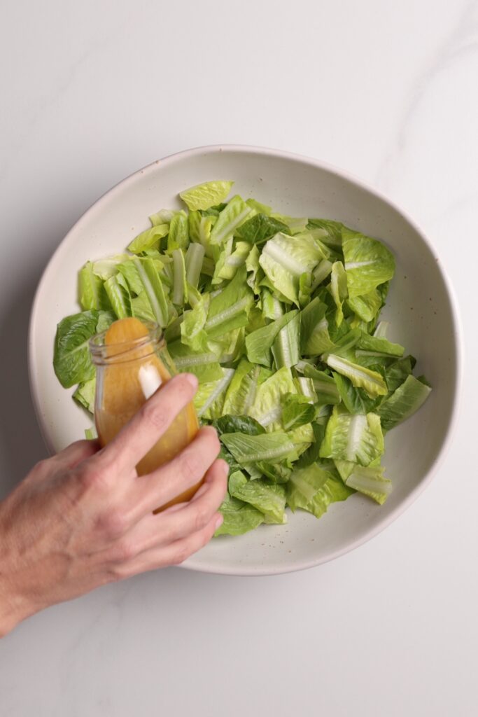 A hand holds a jar of dressing above a bowl filled with chopped romaine lettuce on a white surface.