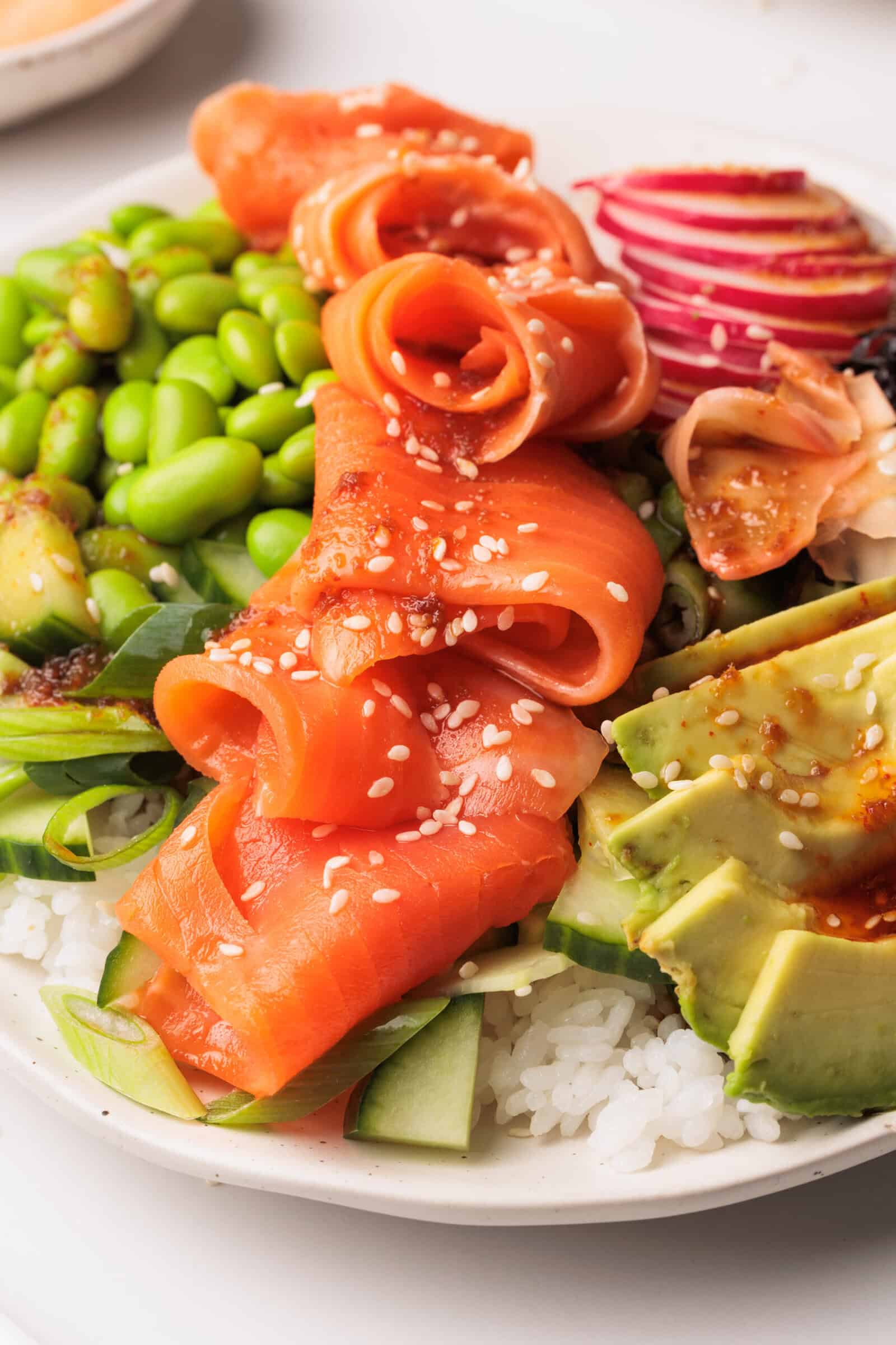 A close-up of a poke bowl topped with slices of smoked salmon, avocado, edamame, cucumber, radish, white rice, sesame seeds, and a drizzle of sauce.