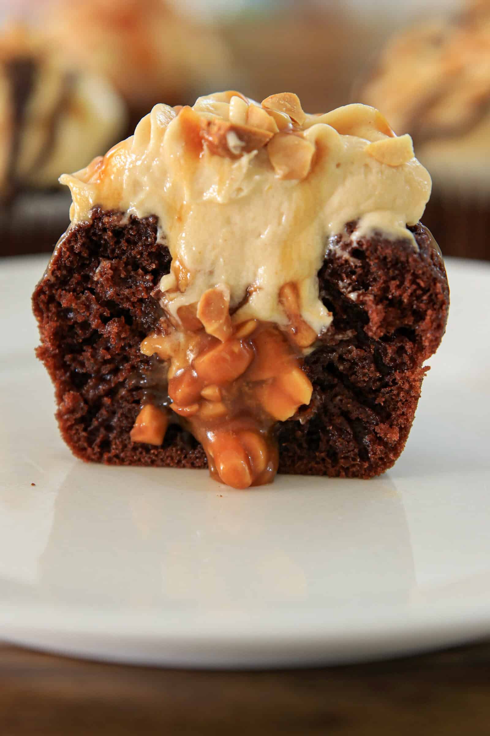 A snickers cupcake cut in half on a white plate, revealing a gooey caramel and nut filling, topped with creamy frosting and chopped nuts.