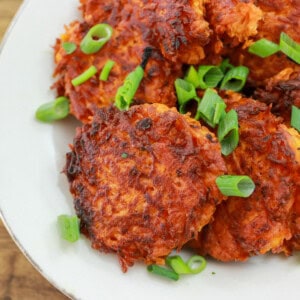 A plate of crispy, golden sweet potato hash browns topped with chopped green onions, served on a white dish.