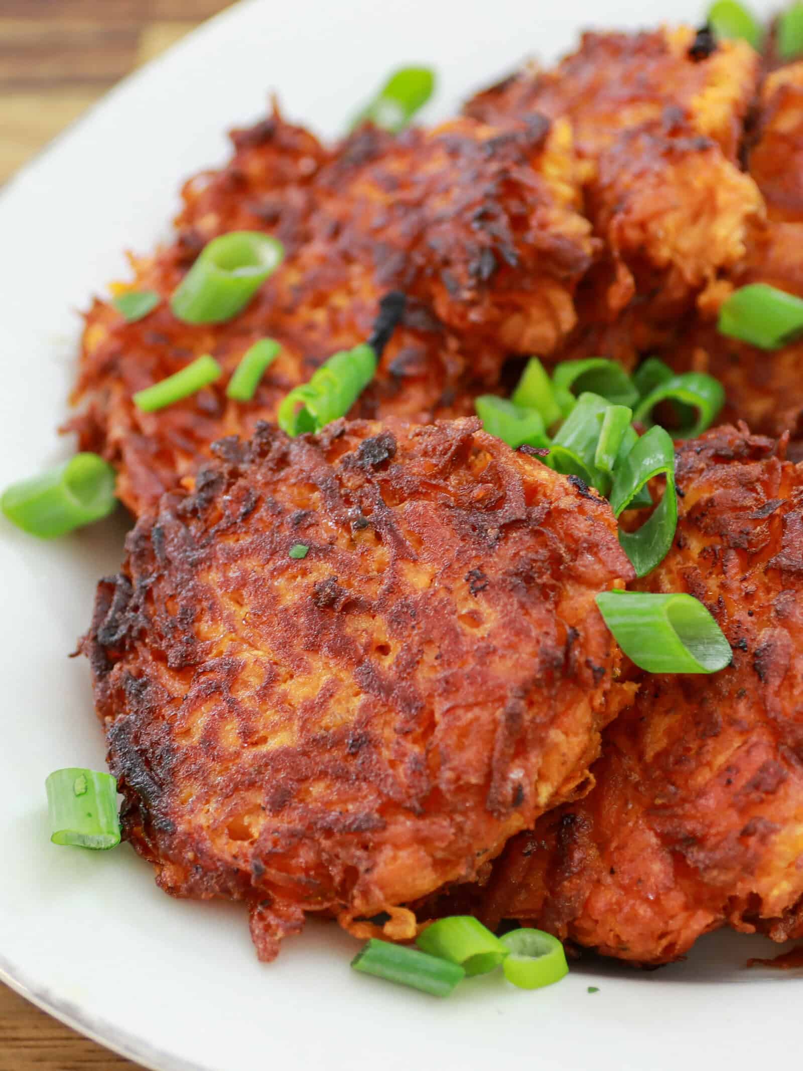 Close-up of crispy, golden-brown sweet potato fritters garnished with chopped green onions, served on a white plate.