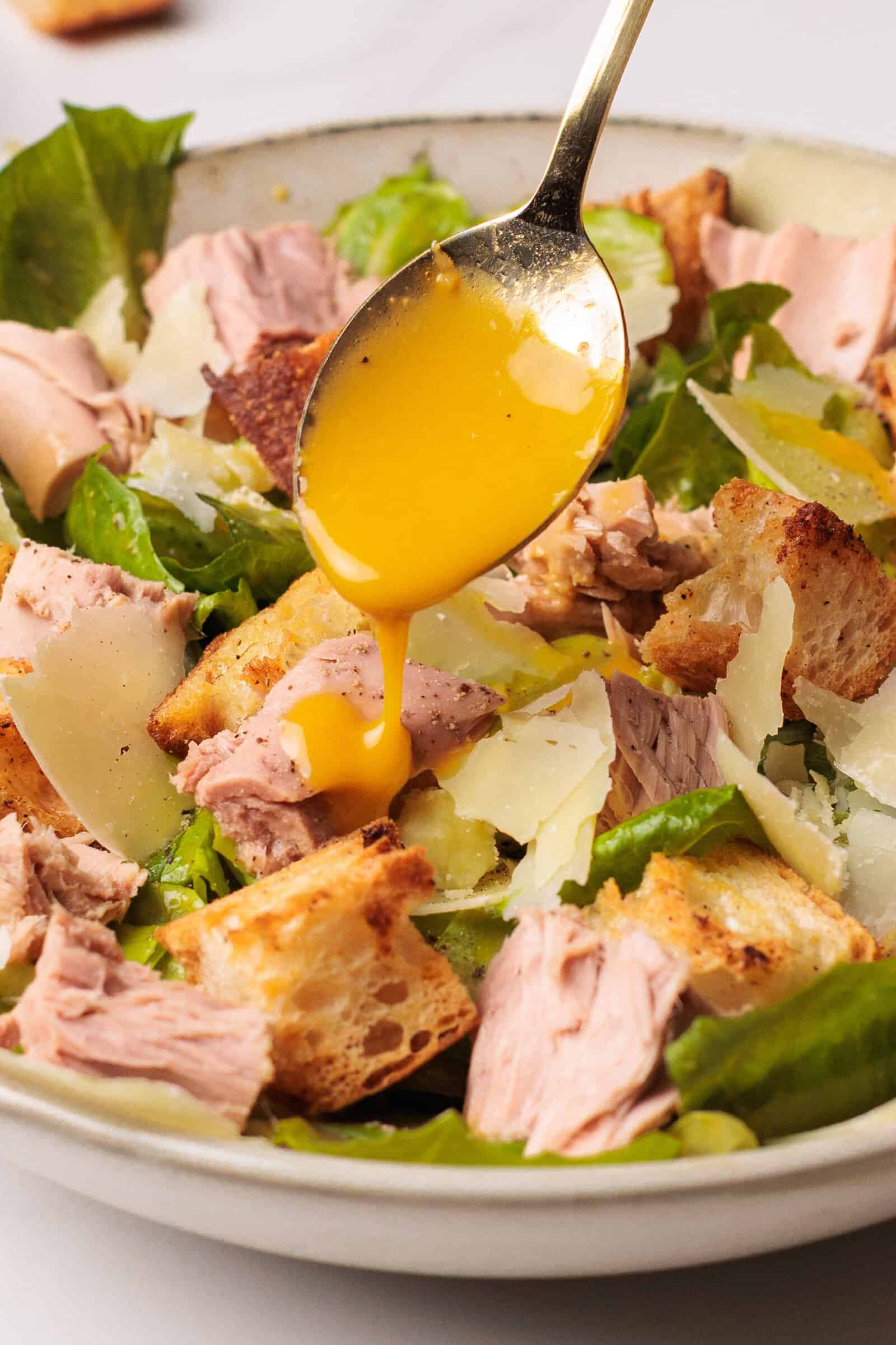 A spoon is drizzling Caesar salad dressing over a bowl of tuna Caesar salad with chunks of tuna, croutons, lettuce, and shaved parmesan cheese.