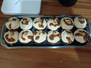 A tray of twelve cupcakes with light frosting and caramel drizzle, each topped with a few peanuts, sits on a wooden surface.