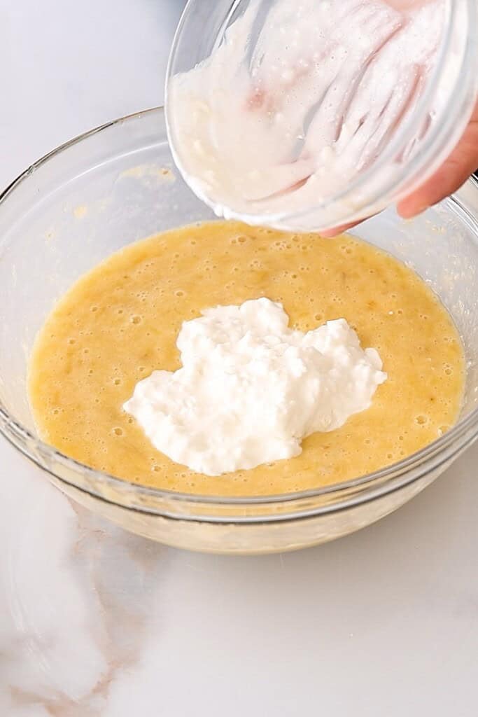 A hand pours a dollop of cottage cheese from a small bowl into a larger bowl containing yellow cake batter on a marble countertop.