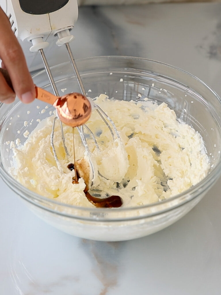 A hand pours vanilla extract from a copper measuring spoon into a bowl of cream cheese frosting, with an electric mixer resting on the bowl on a marble countertop.
