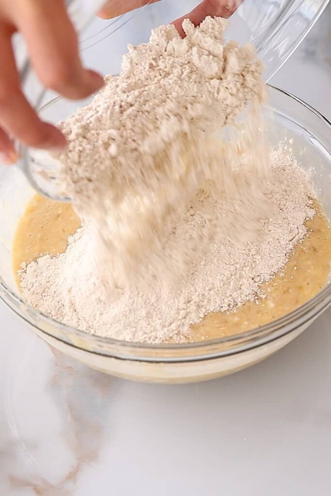 A hand pours a bowl of flour mixture into a larger glass bowl containing a yellow batter, preparing ingredients for baking on a light-colored surface.