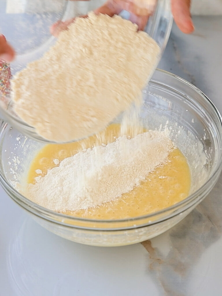 A hand pours flour from a glass bowl into another glass bowl containing a yellow liquid mixture, likely preparing a batter on a marble countertop.