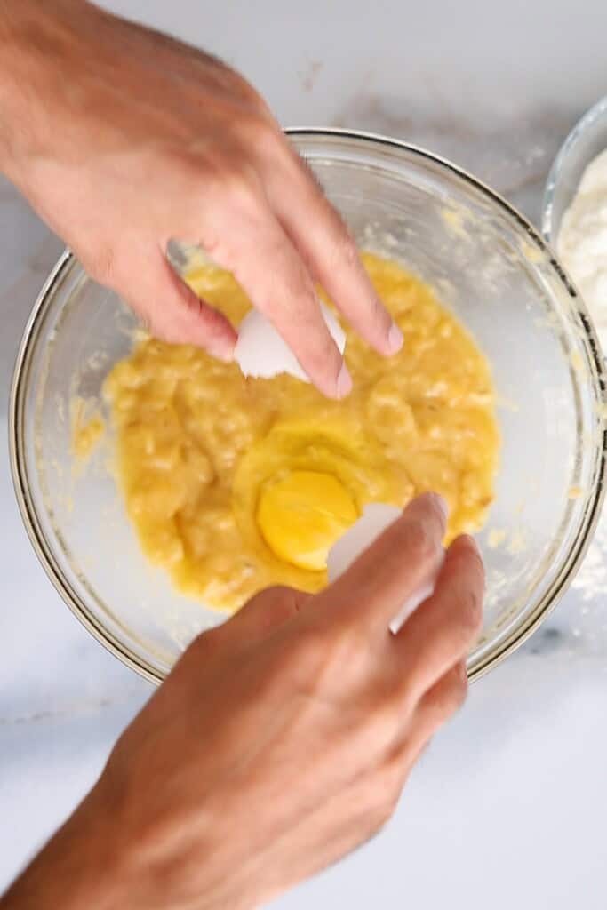 A person cracks an egg into a glass bowl containing mashed bananas and wet ingredients, preparing a mixture for baking.
