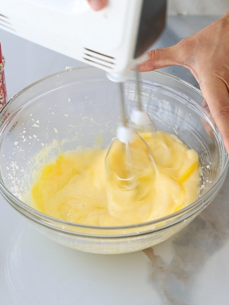 A hand is using an electric hand mixer to blend a yellow batter in a clear glass bowl on a light-colored countertop.