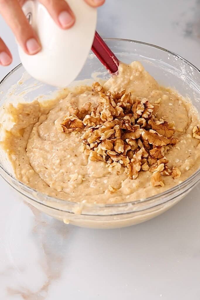 A hand holds a small bowl, pouring walnuts into a mixing bowl filled with thick batter on a light countertop.
