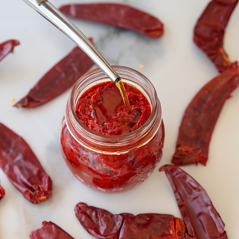 A jar of red chili harissa paste is surrounded by dried red chilies on a white surface. A spoon drizzles oil into the jar, highlighting the vibrant colors and textures of the paste and peppers.