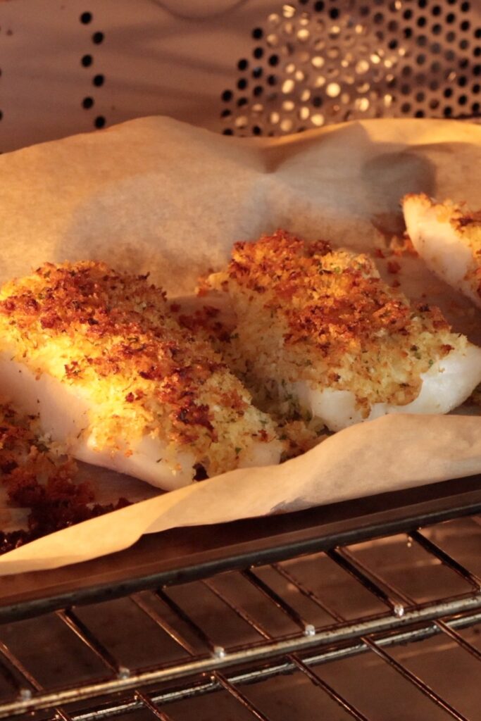 Breaded fish fillets baking in an oven on a parchment-lined tray, with a golden, crispy topping visible under the oven light.