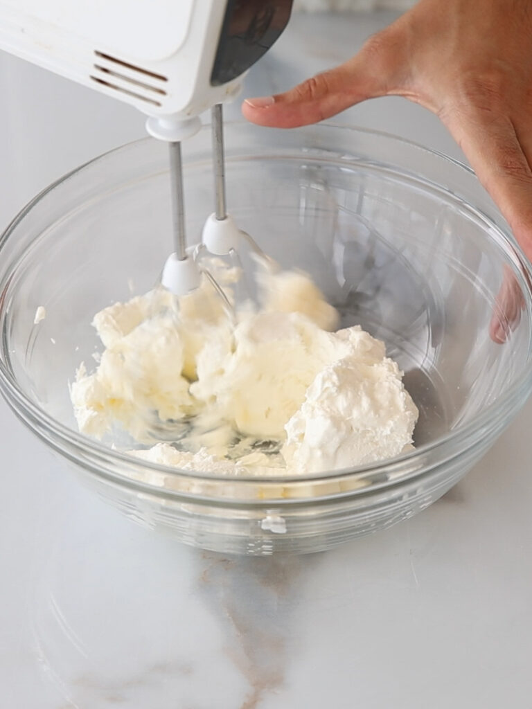 A hand holds an electric mixer, blending butter and powdered sugar in a clear glass bowl on a white countertop.