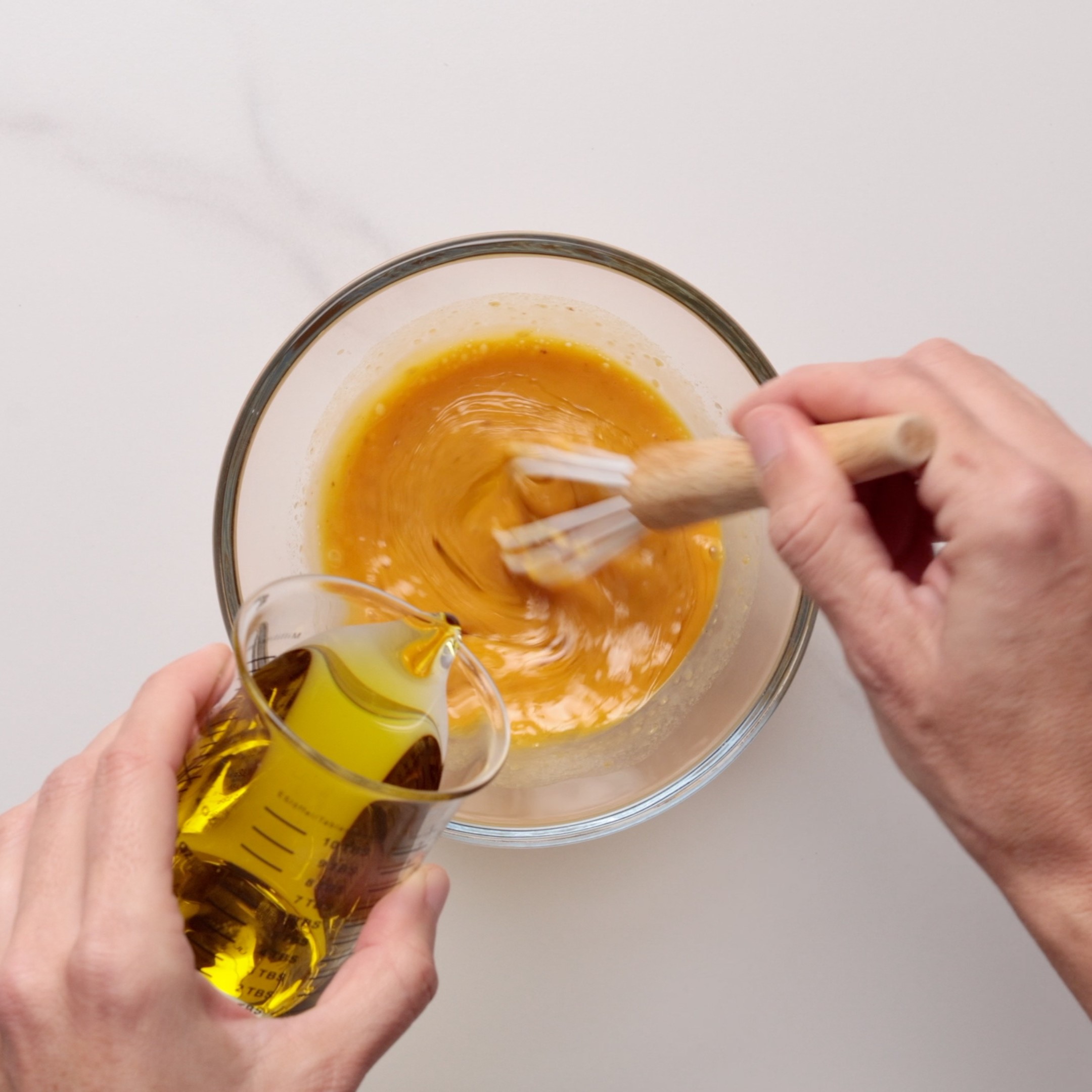A person whisks egg yolks in a glass bowl while pouring oil from a measuring cup onto the mixture, preparing an emulsion on a white countertop.