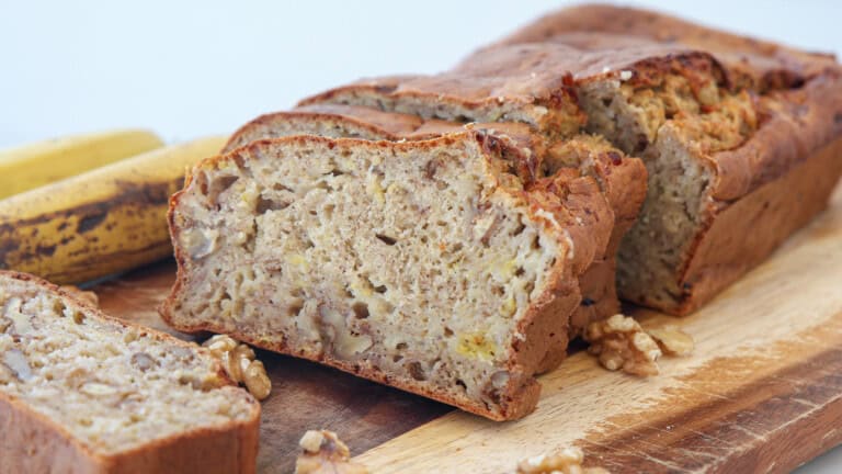 A sliced loaf of cottage cheese banana bread with walnut pieces sits on a wooden board, with whole bananas and walnut halves nearby.